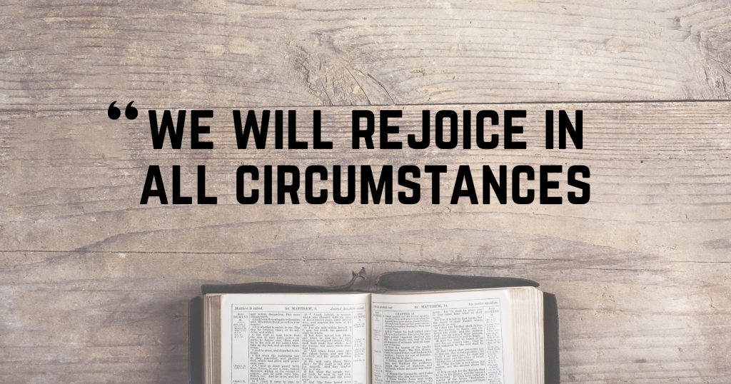 Picture of an open Bible on a wood table with the words, “We will rejoice in all circumstances”