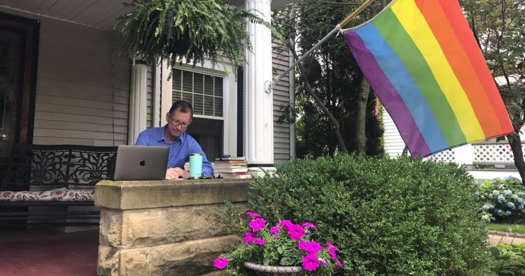 Picture of Rev. Dr. John C. Dorhauer sitting on his porch, writing in a notebook. His laptop, a stack of books, and a coffee cup are nearby. There are many plants, flowers, a bench, and a LGBTQ+ Pride flag around the space.