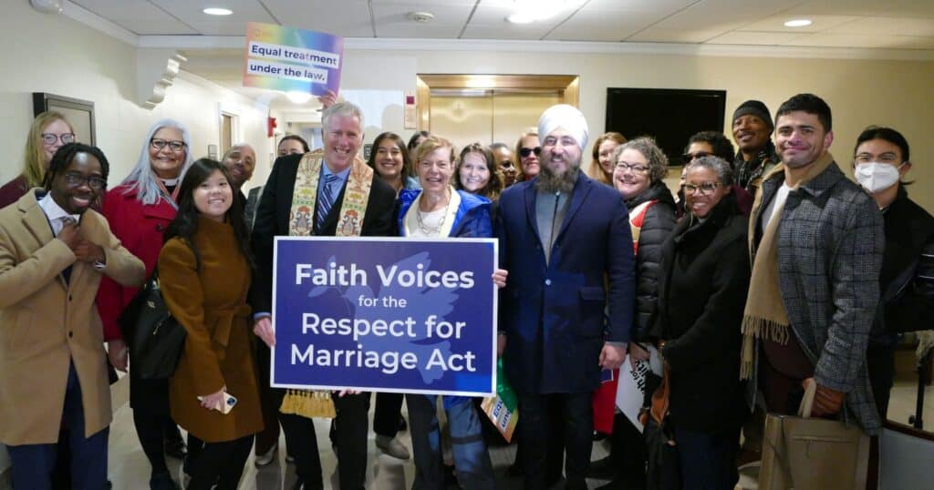 A large group of about two dozen people stand inside to pose for a picture. They are smiling, looking at the camera, and holding a blue sign with a peace dove that says "Faith Voices for the Respect for Marriage Act." The people are wearing coats, and some are wearing religious garments, such as a stole, a clergy collar, or a turban.