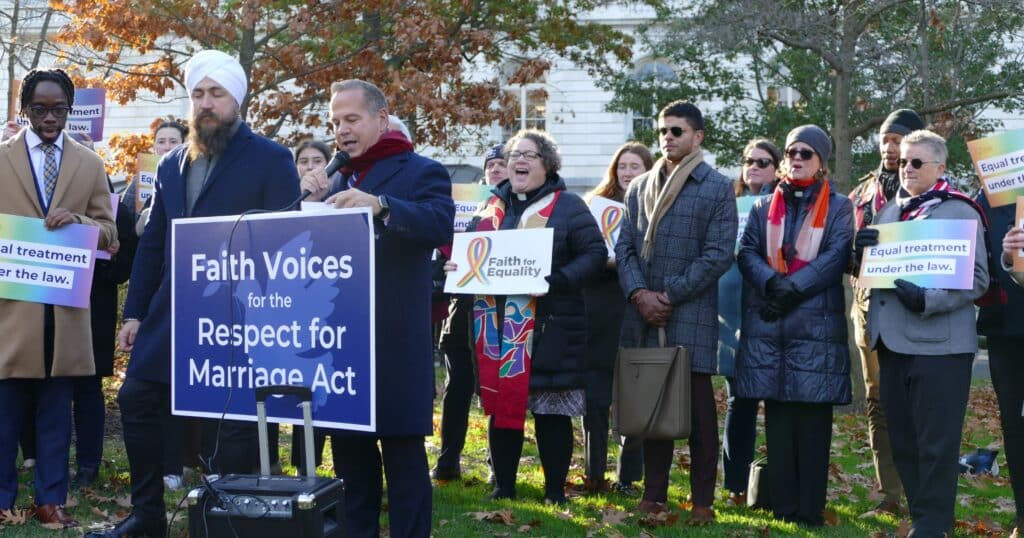 On a sunny autumn day on Capitol Hill, a press conference speaker talks into a microphone and stands behind a sign that says "Faith Voices for the Respect for Marriage Act." He is surrounded by other faith leaders and supporters, many of whom are holding signs and wearing warm coats and scarves. The press conference is taking place outdoors under small trees in front of a white building.