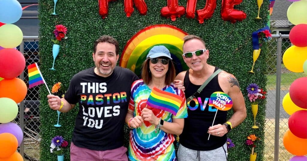 Three people wear rainbow gear in front of a display that says Pride in baloons.