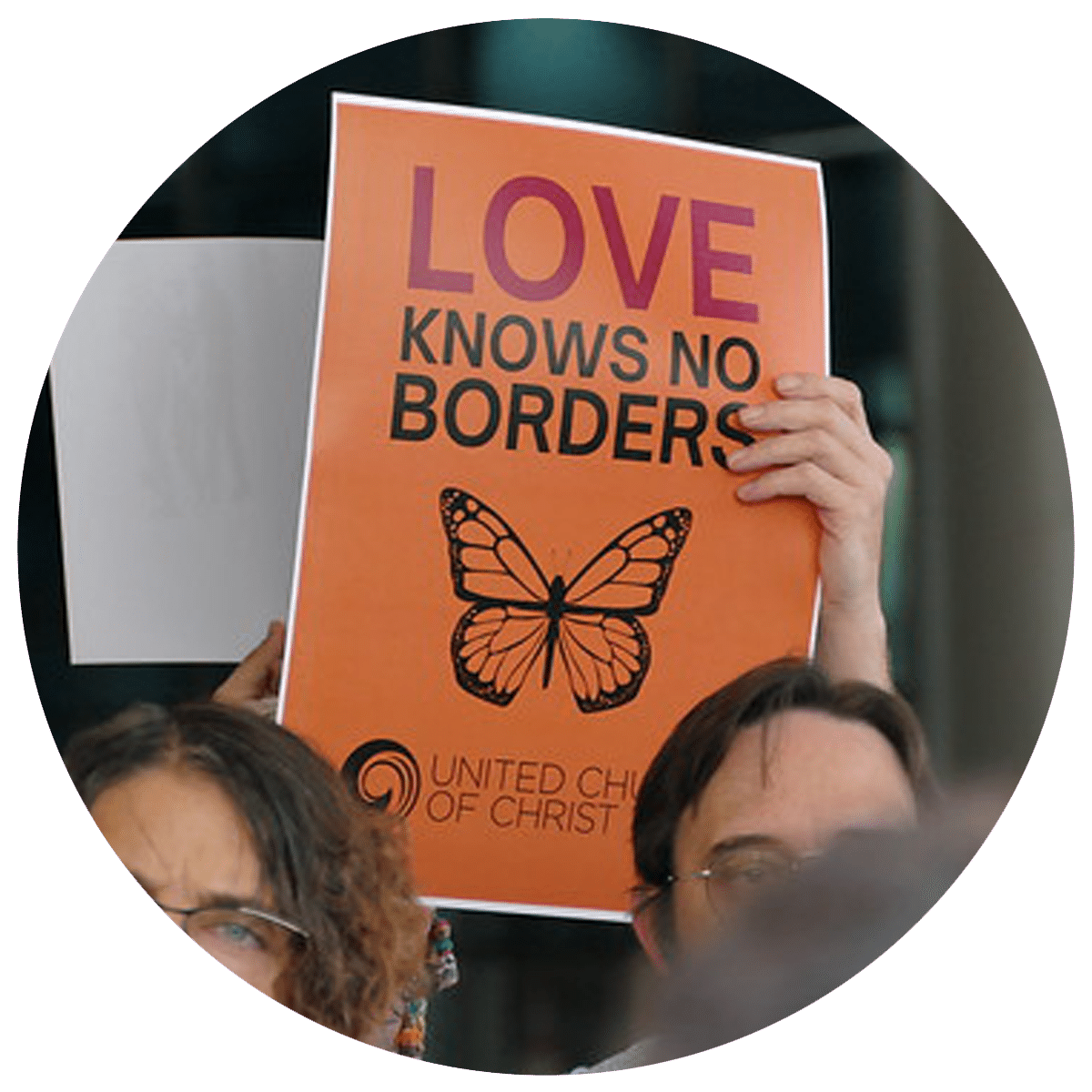 People gather at General Synod 2025 holding signs that read "Love Knows No Borders" on an orange poster with a black outline of a monarch butterfly in the middle.