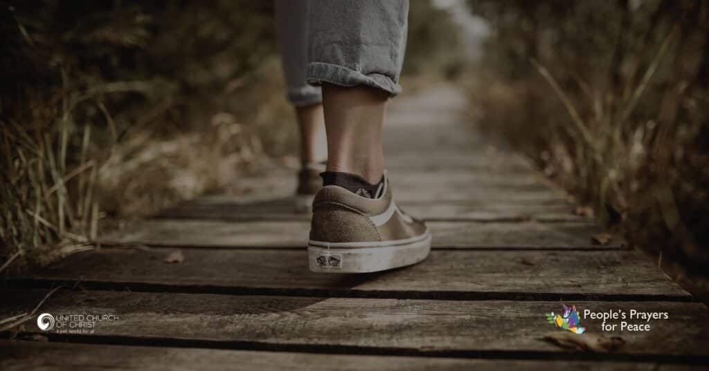 picture in greens and browns depicting an individual walking along a dark brown wooden fence with tall grass on either side. The person is wearing brown tennis shoes and jeans.