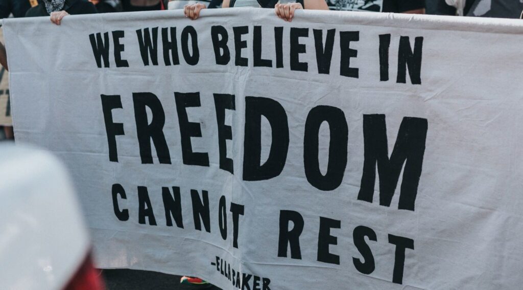 protesters carrying a banner sign that reads We Who Believe in Freedom Cannot Rest - Ella Baker