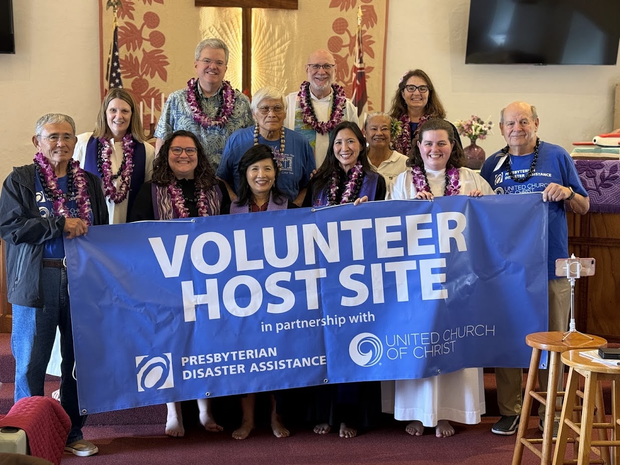 image depicts a group of church volunteers wearing lays holding a blue UCC / Presbyterian Disaster Assistance banner that reads Volunteer Host Site
